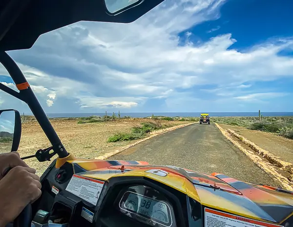 A view from inside an off-road vehicle, possibly a dune buggy or ATV, driving along a coastal road in Aurba with another similar vehicle in the distance. The landscape features arid terrain with scattered vegetation and cacti under a partly cloudy sky, indicating a rugged and adventurous setting during a shore excursion booked in Aruba.