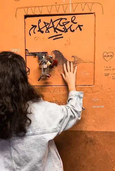 A person looking through the graffiti-covered Little Window on Via Piella, surrounded by padlocks and showcasing a hidden canal in Bologna.