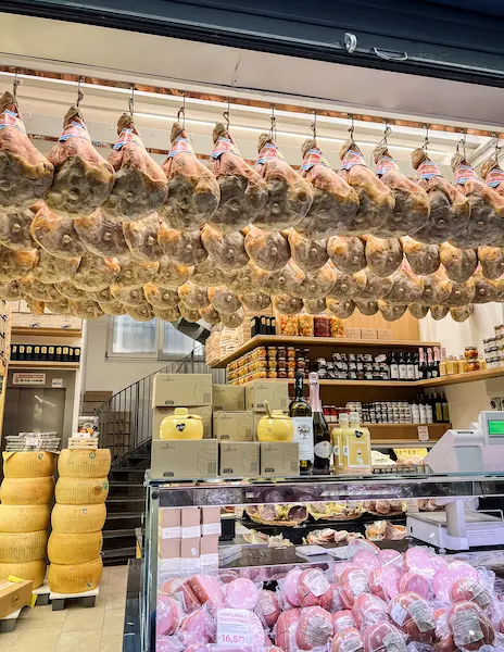 Rows of prosciutto legs hanging in a salumeria in Bologna’s Quadrilatero, surrounded by shelves of cheeses and Italian goods.