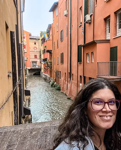 A smiling woman (kathy) taking a selfie with Bologna’s hidden canal in the background, surrounded by vibrant orange buildings