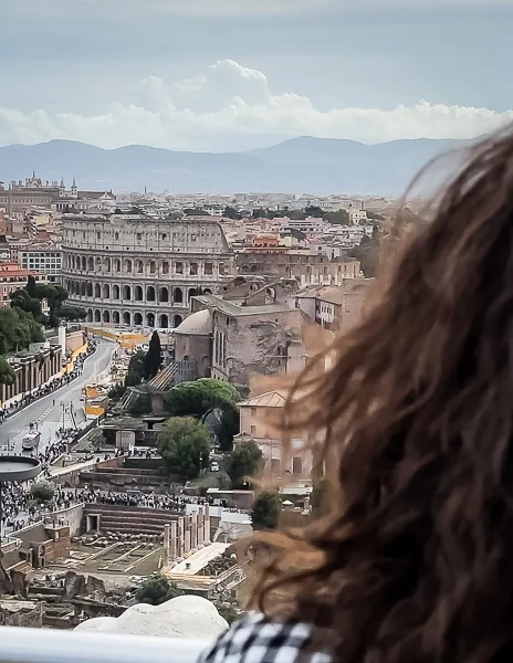 A view of the Colosseum in Rome as seen from the Vittorio Emanuele II Monument. The foreground features part of a person's (kathy) head and hair, while the Colosseum and surrounding cityscape spread out in the background under a cloudy sky.