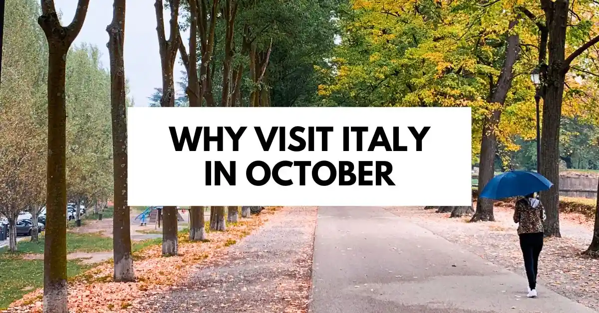 A person walks along a tree-lined path covered in autumn leaves, holding a blue umbrella. The image captures the essence of fall in Italy with vibrant foliage, highlighting the appeal of visiting in October. The text overlay reads "Why Visit Italy in October."