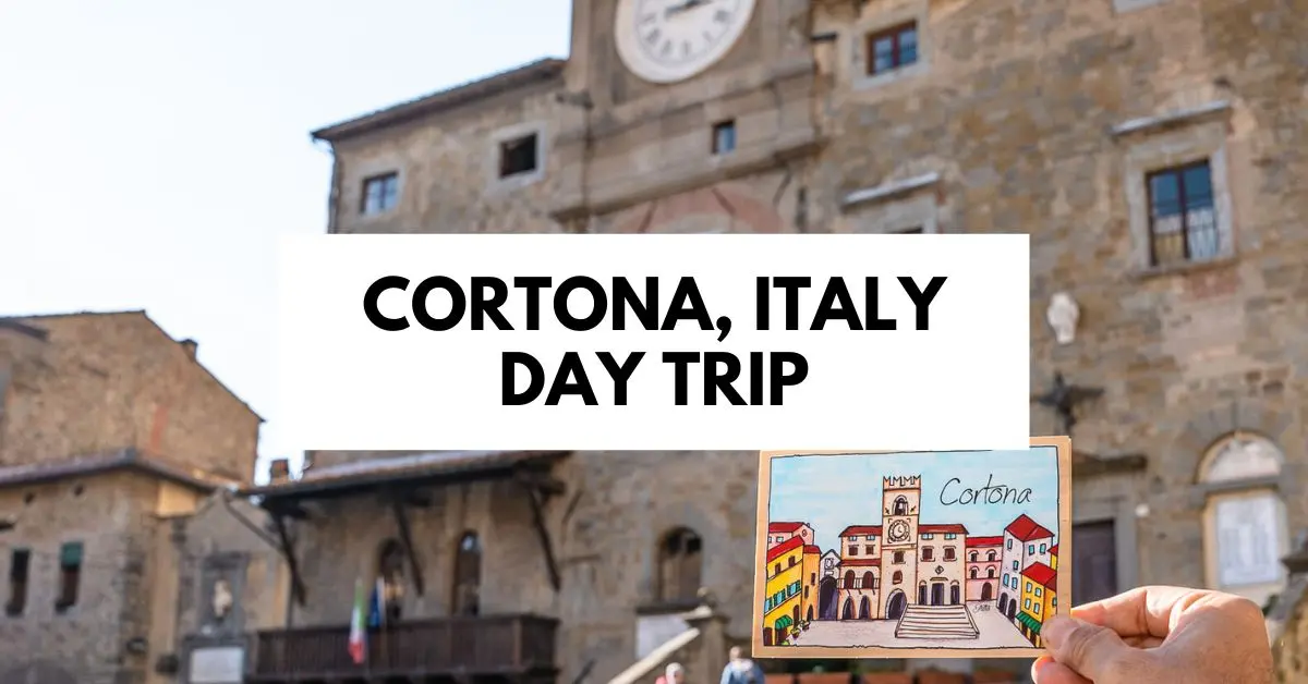 A hand holding a colorful postcard illustration of the main square in Cortona, Italy, with the historic town square and clock tower in the background. The text overlay reads "Cortona, Italy Day Trip," highlighting this charming Tuscan destination.