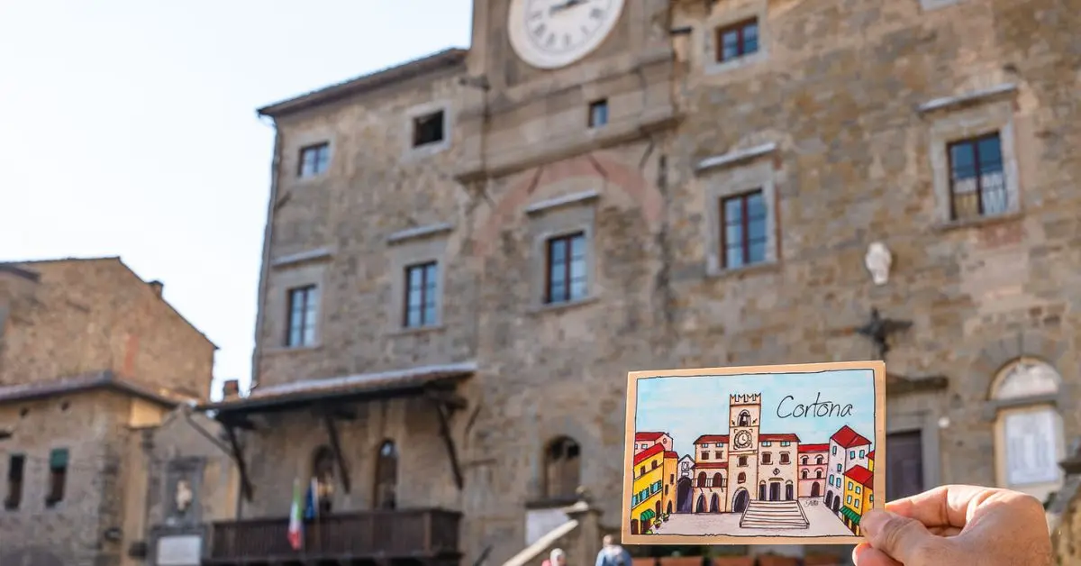 A hand holding a colorful postcard of Cortona in front of the iconic Palazzo Comunale in Piazza della Repubblica, perfectly capturing the charm of one day in Cortona.