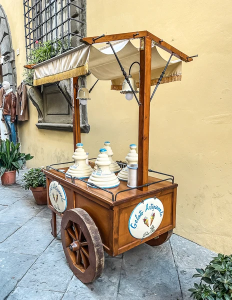 A vintage wooden gelato cart with a canopy, labeled "Gelato Artigianale," parked on a cobblestone street in Cortona, Italy, adding charm to the setting.