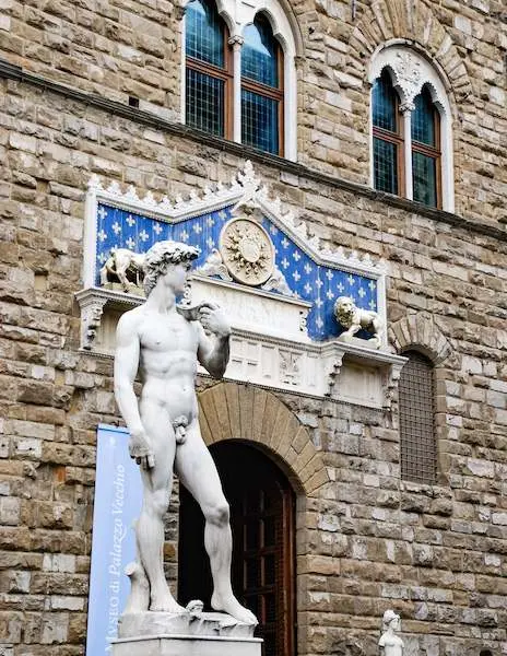 A marble replica of Michelangelo's David statue standing in front of the Palazzo Vecchio at Piazza della Signoria in Florence, Italy, with intricate architectural details and blue accents in the background.