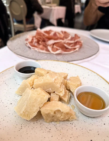 A plate of aged Parmigiano Reggiano cheese, cut into chunks, accompanied by small bowls of balsamic vinegar and honey. In the background, a plate of thinly sliced Prosciutto di Parma completes the setting, highlighting iconic foods from Emilia-Romagna, Italy.