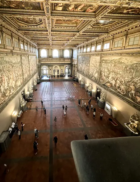 The grand Sala dei Cinquecento inside Palazzo Vecchio, adorned with detailed frescoes, statues, and a high coffered ceiling, viewed from above.