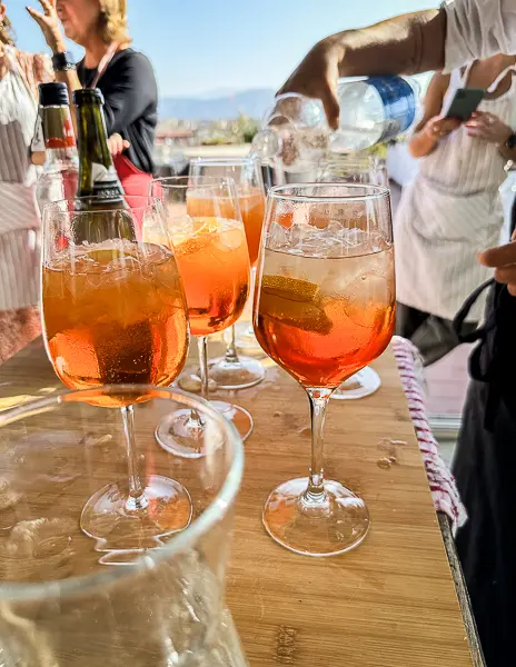 Glasses of Aperol Spritz on a wooden table during a cooking class in Florence, with participants in aprons gathered in the background.