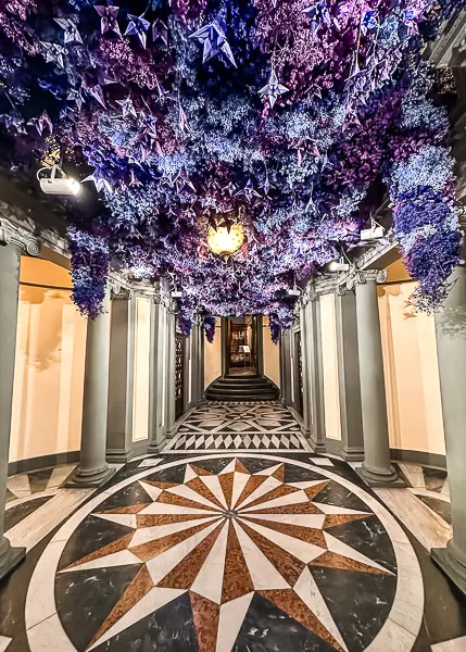 The lobby of the Farmaceutica di Santa Maria Novella in Florence, adorned with vibrant purple flowers hanging from the ceiling and a geometric marble floor design. , adorned with vibrant purple flowers hanging from the ceiling and a geometric marble floor design.