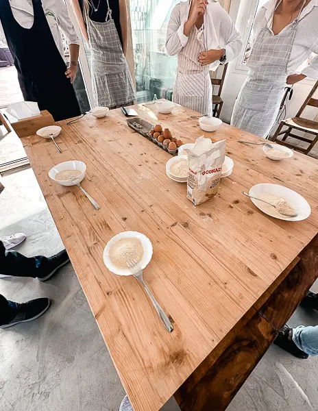 A table set up for a cooking class in Florence, Italy, with bowls of flour, eggs, and utensils ready for making pasta. Participants in aprons stand around the table.