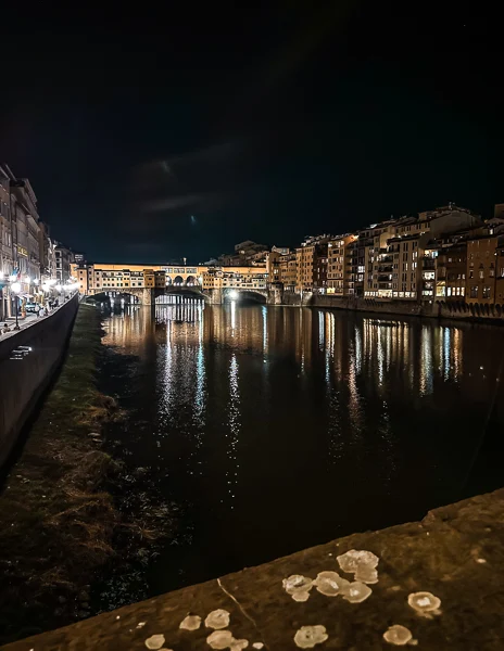 A nighttime view of the Ponte Vecchio in Florence, beautifully lit with its reflection shimmering on the calm waters of the Arno River.