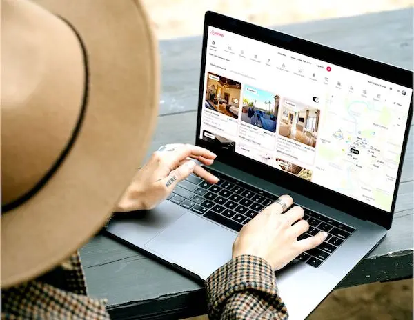 a person using a laptop to browse Airbnb listings, possibly researching for their next travel accommodation. The laptop screen displays various Airbnb options with images, descriptions, and a map. 