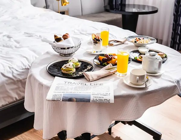 a breakfast setup in a cozy bedroom, likely delivered as room service. A small round table covered with a white tablecloth holds an assortment of breakfast items, including a teapot, cups, orange juice, pastries, and a newspaper.