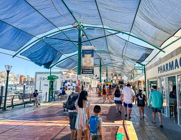 A busy walkway at Cabo San Lucas Cruise Port, shaded by a large blue canopy. People are strolling along, some stopping at the various shops on the right, which include a pharmacy. The scene captures a sunny day, with clear skies visible beyond the canopy.