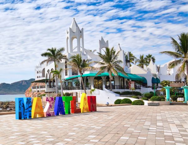 The colorful Mazatlán sign in the Golden Zone with a white architectural building and palm trees in the background.