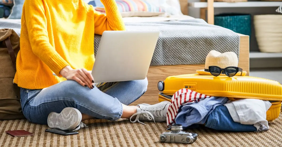 featured blog image of a person sitting on the floor with a laptop, next to a packed suitcase, camera, and travel essentials, thinking about how to use these travel tips for beginners.