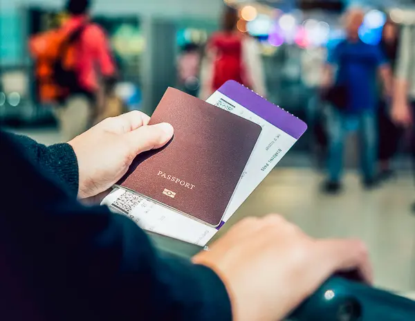 A close-up of a person holding a passport and a boarding pass at an airport, with blurred travelers and luggage in the background.