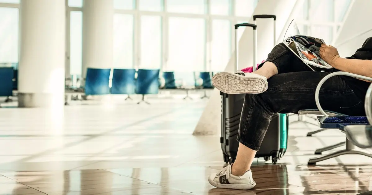 A traveler sitting in an airport with their feet up on a chair, reading a magazine, with luggage nearby. This image represents common travel mistakes to avoid.