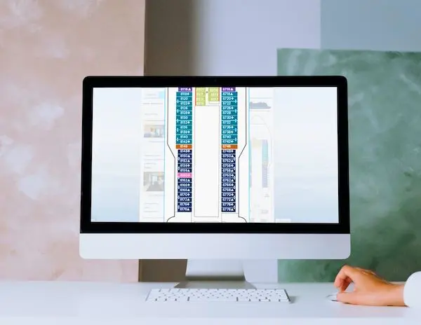 image shows a desktop computer screen displaying a cruise ship deck plan. The screen provides a detailed layout of the ship's cabins, color-coded to indicate different categories or types. A person is visible at the desk, partially interacting with the computer