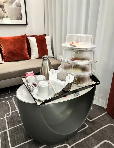 Room service tray with stacked clear containers of food, carafe of coffee, cups, and utensils, placed on a small round table in a cozy room with a red-accented sofa.