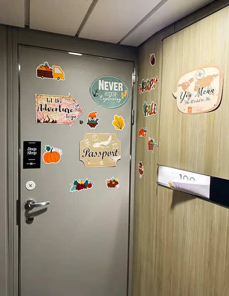 a cruise cabin door decorated with various colorful magnets and stickers. The decorations include travel-themed slogans like "Never stop exploring," "Passport," and other playful graphics such as a camper van, fruits, and autumn leaves. 