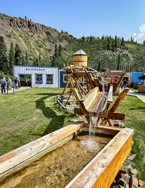 A gold panning station at Wild Adventure Yukon with a wooden water wheel and sluice, surrounded by lush grass and set against a backdrop of rustic buildings and hills.