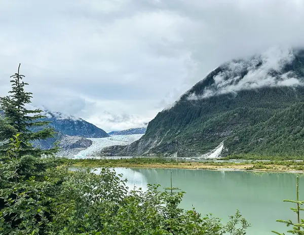 Vew of Mendenhall Glacier from photo point trail in Juneau, Alaska, with a serene turquoise lake in the foreground, flanked by lush greenery and framed by mist-covered mountains.