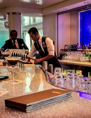 An image of two bartenders preparing drinks at a cruise ship bar, with a bar menu on the glittery counter and a row of espresso martinis in the background, under a blue-lit shelf.