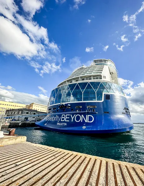 view of the aft of the Celebrity Beyond cruise ship, docked in a Naples cruise port. The ship’s name is prominently displayed against the vibrant blue hull, with its home port of Valletta also visible. The ship's modern design is accentuated by the expansive glass-encased structure towards the stern, set against a backdrop of a vivid blue sky with scattered clouds. 