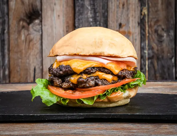 A cheeseburger with double beef patties, cheese, lettuce, and tomato, on a bun, presented on a black slate surface with a rustic wooden backdrop.