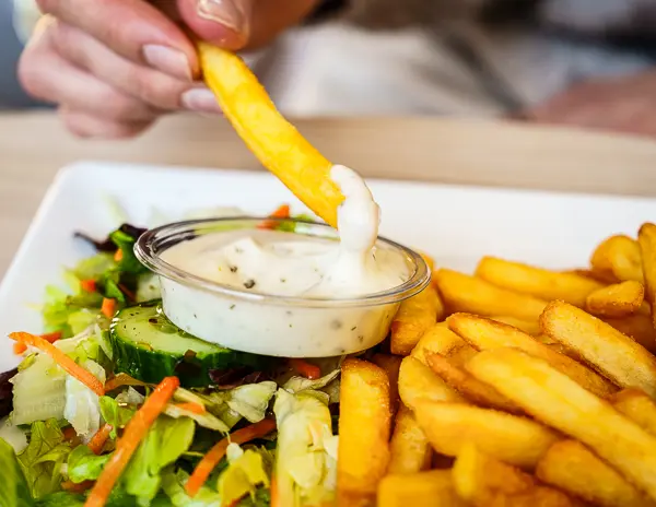 A hand dipping a French fry into a small container of ranch dressing, with a side of salad and more fries on a white plate.