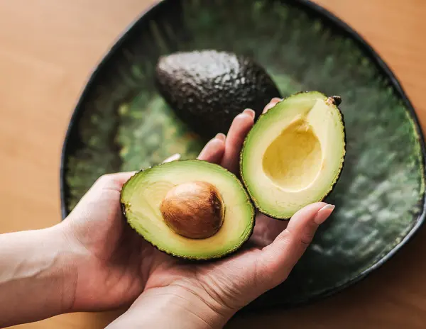 A person's hands holding a halved Hass avocado over a dark green plate, with another whole avocado beside it.