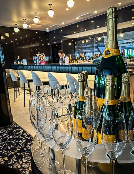  a cruise ship champagne bar, foregrounded by a selection of sparkling wine bottles and elegant flutes ready for service. In the softly lit background, a waiter is seen attending to the bar, contributing to the sophisticated and luxurious atmosphere of the setting