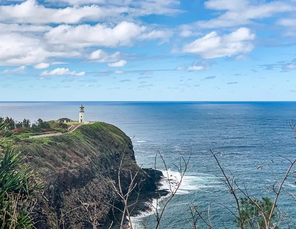 Kilauea lighthouse perched on a steep, grassy cliff overlooking the ocean, with waves crashing against the rocky shore below, under a partly cloudy sky