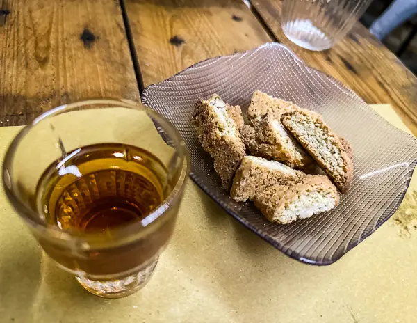 A glass of Vin Santo with a side of Cantucci biscuits served on a textured glass plate, set on a wooden table.