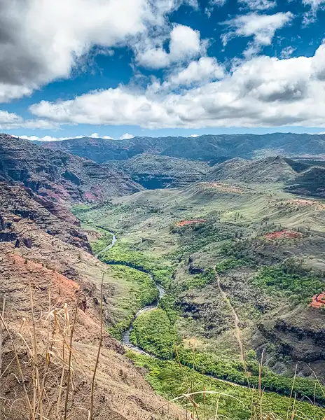 A sweeping view of Waimea Canyon, showcasing rugged terrain, deep valleys, and a meandering river, under a dynamic sky with scattered clouds.