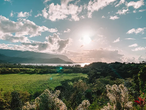 Sunset over lush green fields with a view of the sea and distant mountains in Princeville, Kauai, under a vibrant sky scattered with clouds.
