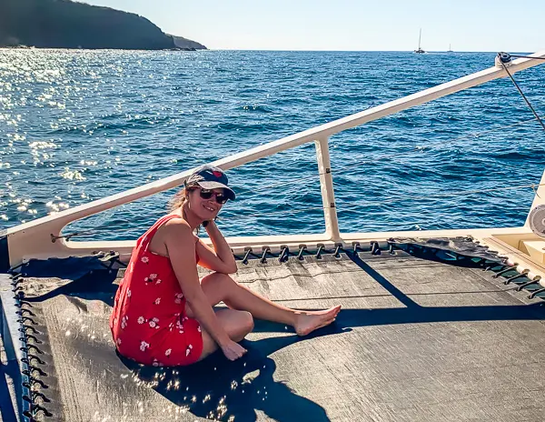 kathy sitting on the deck of a yacht, wearing a red dress with floral patterns and a baseball cap. She is smiling at the camera, with the sparkling blue ocean and a distant hill in the background, capturing a sunny day on the sea.