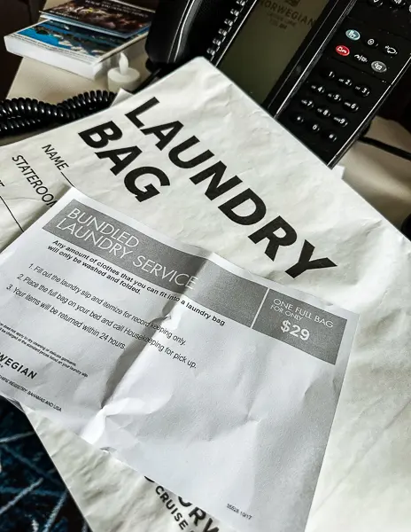 A laundry bag from a cruise line, specifically labeled for a bundled laundry service. It includes pricing information, noting that one full bag costs $29. The bag is placed on a table next to a cruise ship telephone and other room documents, suggesting it's ready for use by the cabin occupant.