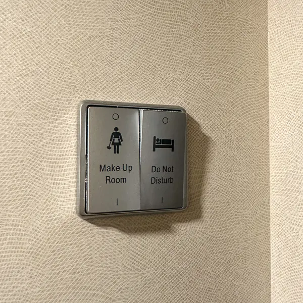 Two buttons on textured wall: "Make Up Room" and "Do Not Disturb." The "Make Up Room" button is marked with an icon of a figure and a broom, signaling housekeeping to tidy the room. The "Do Not Disturb" button shows a bed icon, indicating that the occupants do not wish to be disturbed