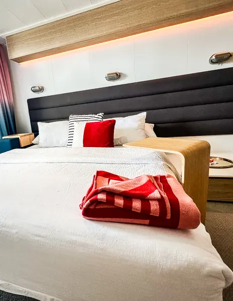 a neatly arranged cruise ship cabin bed with a large headboard and stylish bedding. A red throw blanket is neatly folded at the foot of the bed, adding a pop of color to the room's decor. 