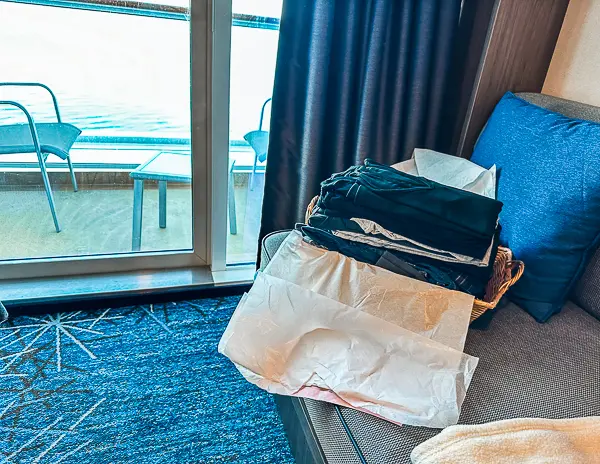 a basket of neatly folded laundry, including various clothing items, placed on a couch inside a stateroom on a Norwegian Cruise Line ship. The room features a balcony visible through the sliding glass door, suggesting a pleasant and comfortable onboard accommodation.