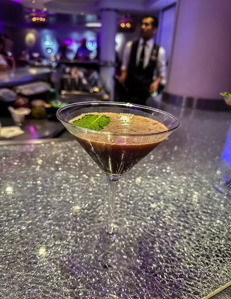 An image of a chocolate martini garnished with mint, served in a martini glass on a glittery bar counter. The setting features a softly lit bar with a bartender in the background