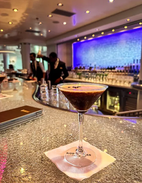 A photo of an espresso martini served on a glittery bar counter. The cocktail is presented in a martini glass, set on a napkin labeled "NORWEGIAN". In the background, the bar displays a luminous blue light and bartenders are visible, attending to other tasks.