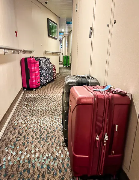 a corridor on a Norwegian Cruise Line ship, lined with various suitcases. The carpet in the hallway features a pattern with fish, which are used as navigational aids, pointing towards the front of the ship. This is a practical design element intended to help passengers orient themselves as they move through the ship's corridors.