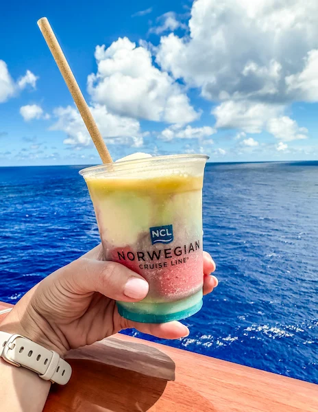 An image of a hand holding a Miami Vice cocktail in a "NORWEGIAN CRUISE LINE" branded cup, with distinct yellow, blue, and red layers. The background features a vibrant blue ocean and sky, viewed from a cruise ship.