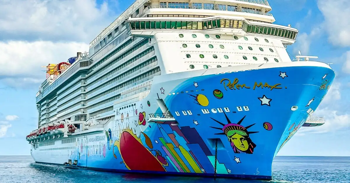 featured image without text features the Norwegian Breakaway cruise ship, highlighted by its colorful hull artwork designed by Peter Max. The ship, shown in a side view, displays the iconic Statue of Liberty design amidst a playful and vivid background of stars and planets, set against the backdrop of a clear blue sky and ocean.
