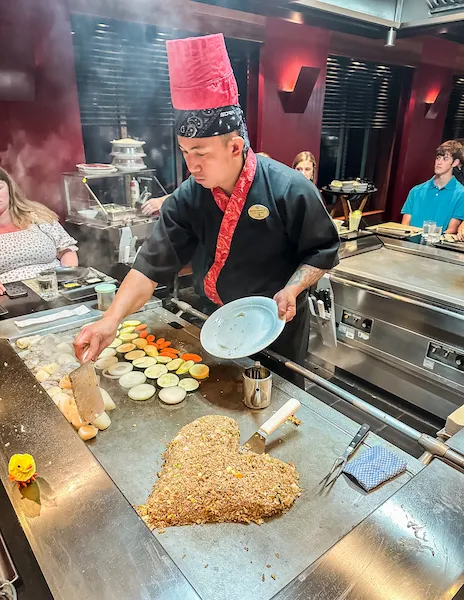 a Teppanyaki chef preparing food on a grill, with shaped fried rice and vegetables in a lively, interactive dining environment.