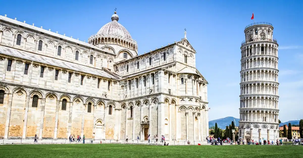 featured blog image without text is a wide-angle shot showcasing both the Pisa Cathedral and the Leaning Tower of Pisa with tourists visiting. The entire scene is set against a clear blue sky.
